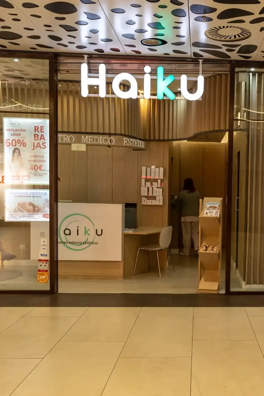 Entrada de la tienda Haiku con logotipo y una persona dentro. Interior de madera clara, puertas de cristal y un cartel en el techo.