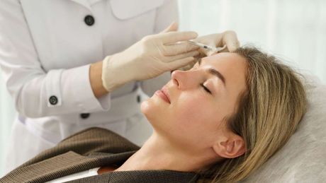 Persona recibiendo una inyección facial con una jeringa en la mano, enguantada en rosa. El paciente lleva una redecilla para el pelo.