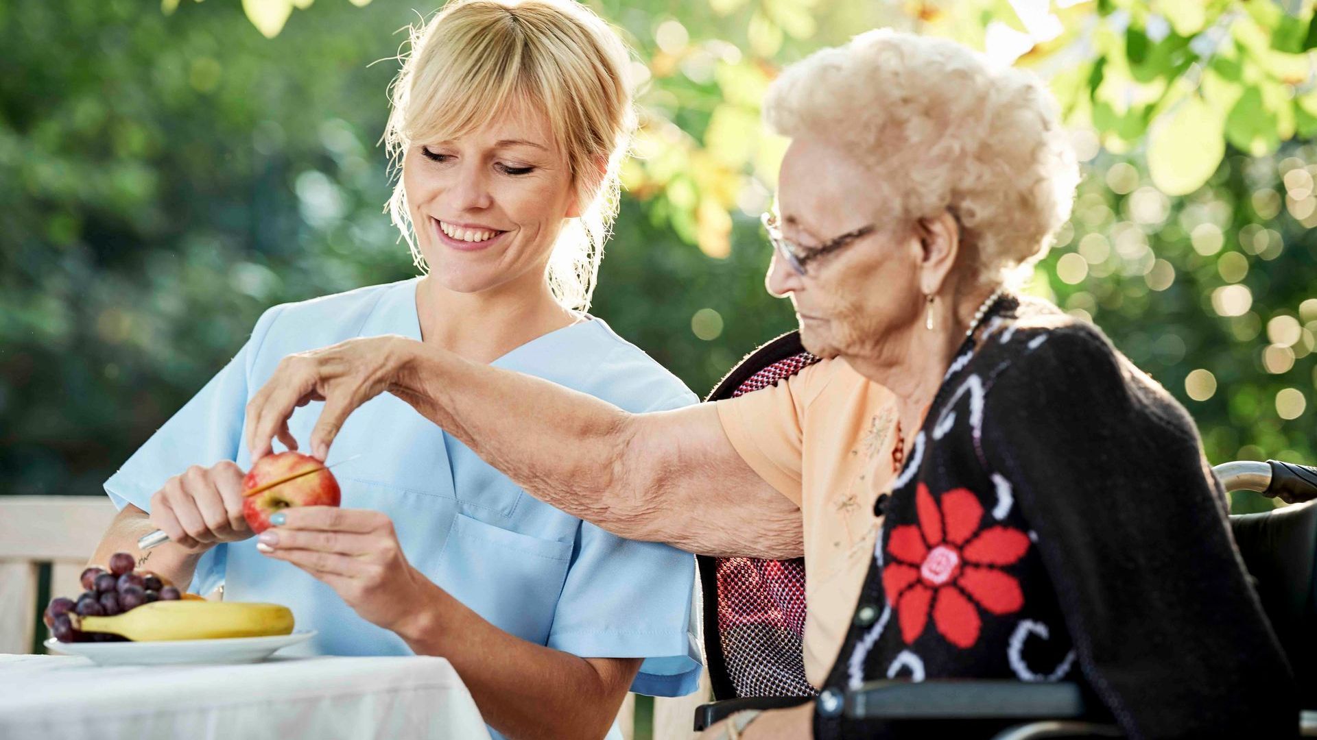 Eine Krankenschwester gibt einer älteren Frau im Rollstuhl einen Apfel.