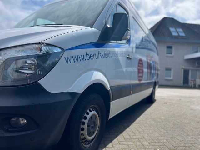 Weißer Lieferwagen mit blauem Streifen und Firmenlogo, geparkt auf einer gepflasterten Fläche in der Nähe eines Gebäudes.