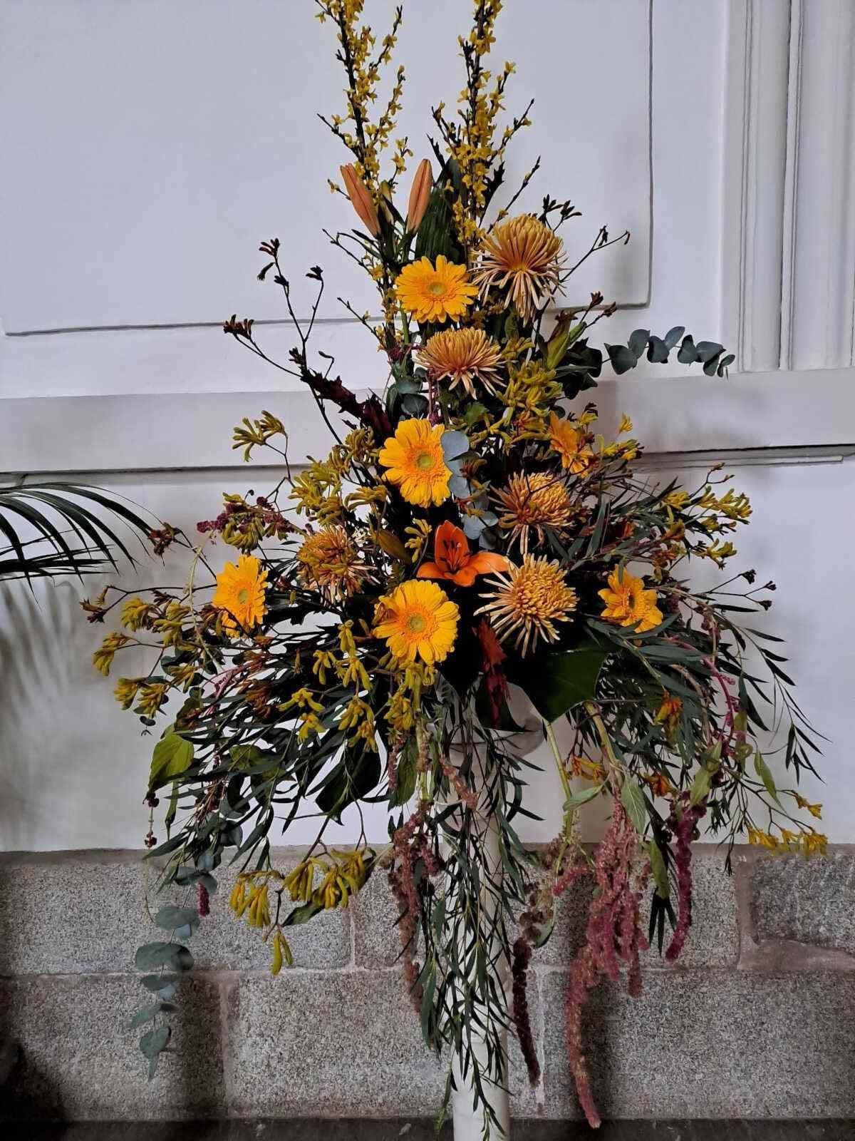 Un jarrón lleno de flores amarillas y vegetación está sobre una mesa.