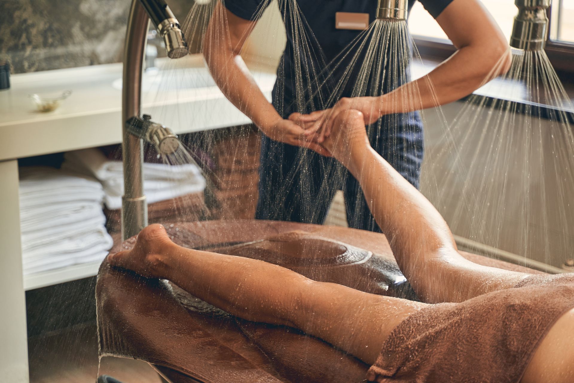 Personne recevant un massage des jambes dans une douche ou un spa, avec de l’eau projetée à proximité.