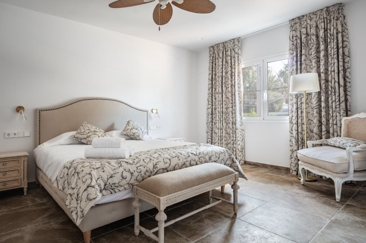 Un dormitorio con una cama, silla, banco y ventilador de techo.