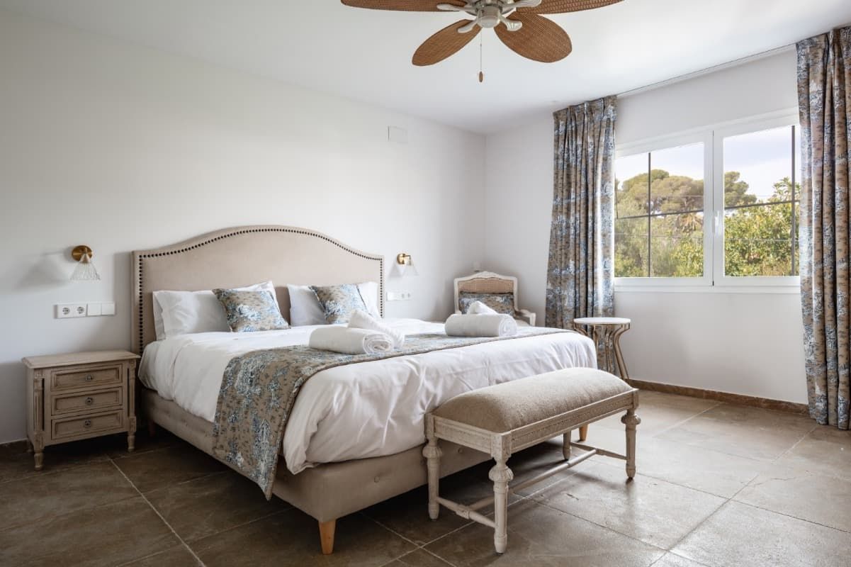 Un dormitorio con una cama king size, mesita de noche, banco y ventilador de techo.