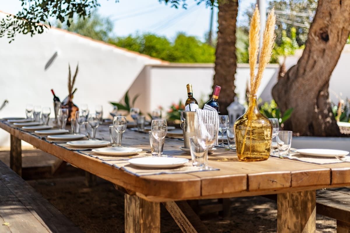 Una larga mesa de madera con platos, vasos y botellas sobre ella.