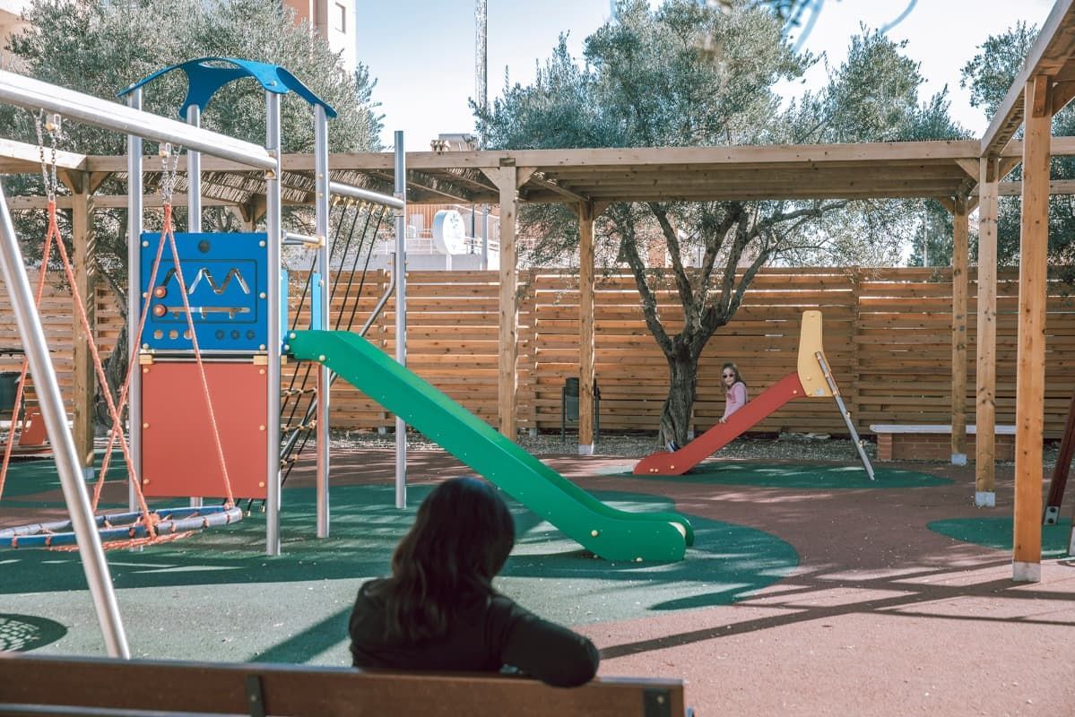 Una persona está sentada en un banco en un patio de recreo mirando un tobogán.