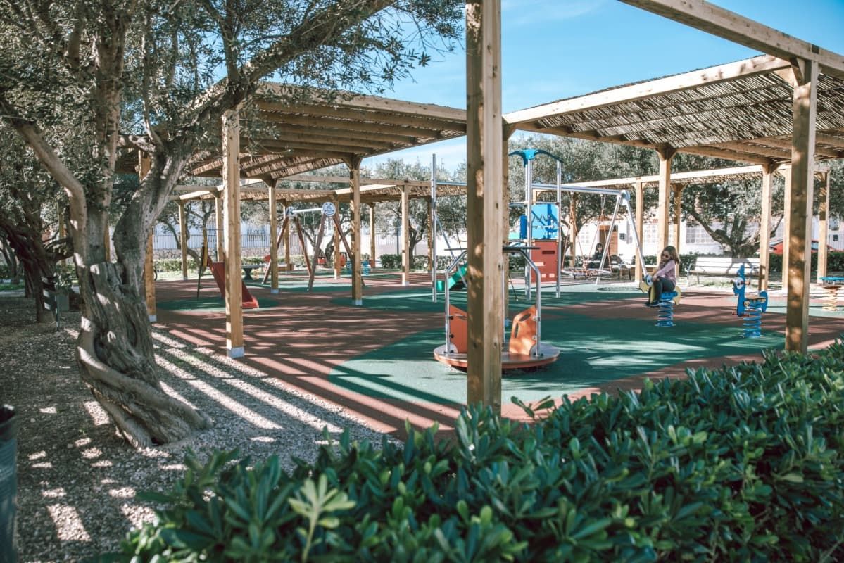 Un patio de juegos en un parque con una zona cubierta y árboles al fondo.