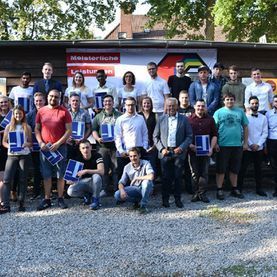 Eine große Gruppe posiert im Freien vor einem Gebäude, einige halten blaue Mappen, über ihnen Bäume.