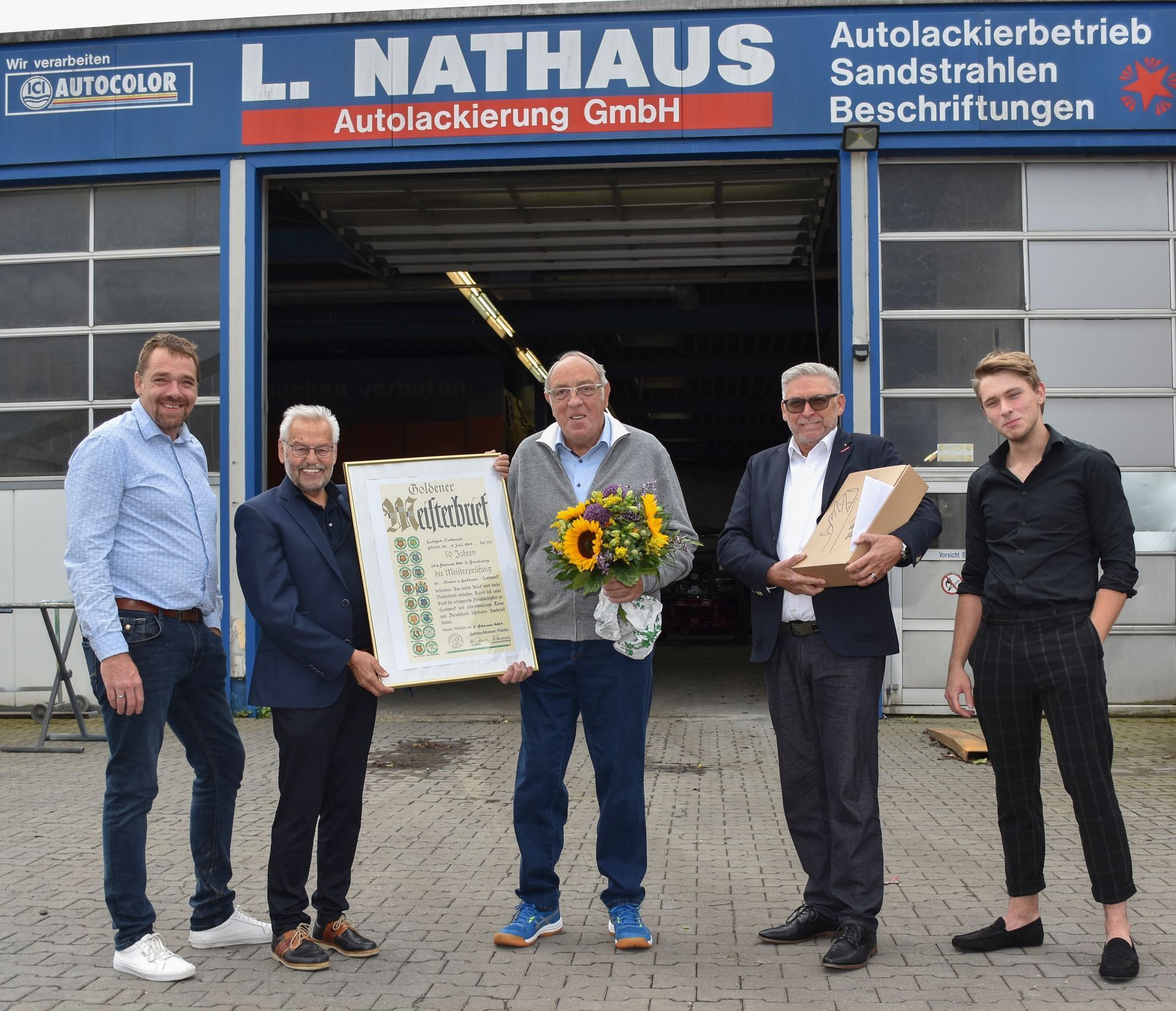 Fünf Personen posieren vor einer Autolackiererei und halten eine gerahmte Urkunde und einen Blumenstrauß in den Händen.