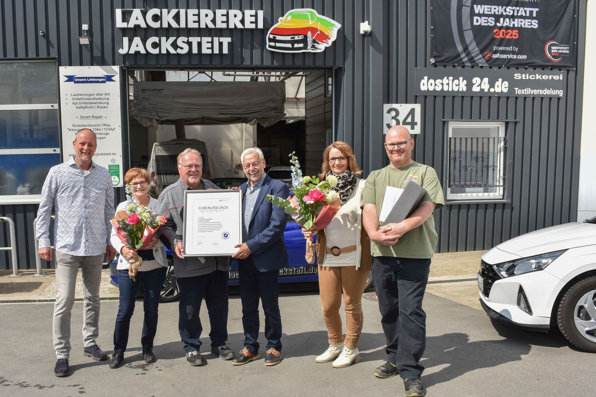 Fünf Personen posieren vor einer Werkstatt, sie halten Blumen und eine gerahmte Urkunde.