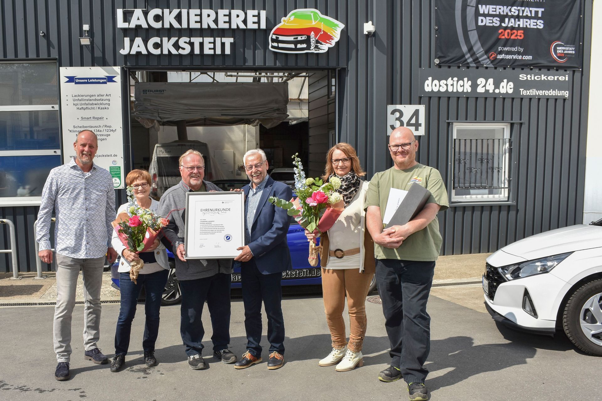 Eine Gruppe posiert vor einer Werkstatt, hält Blumen und eine gerahmte Urkunde in den Händen; rechts ist ein Auto zu sehen.
