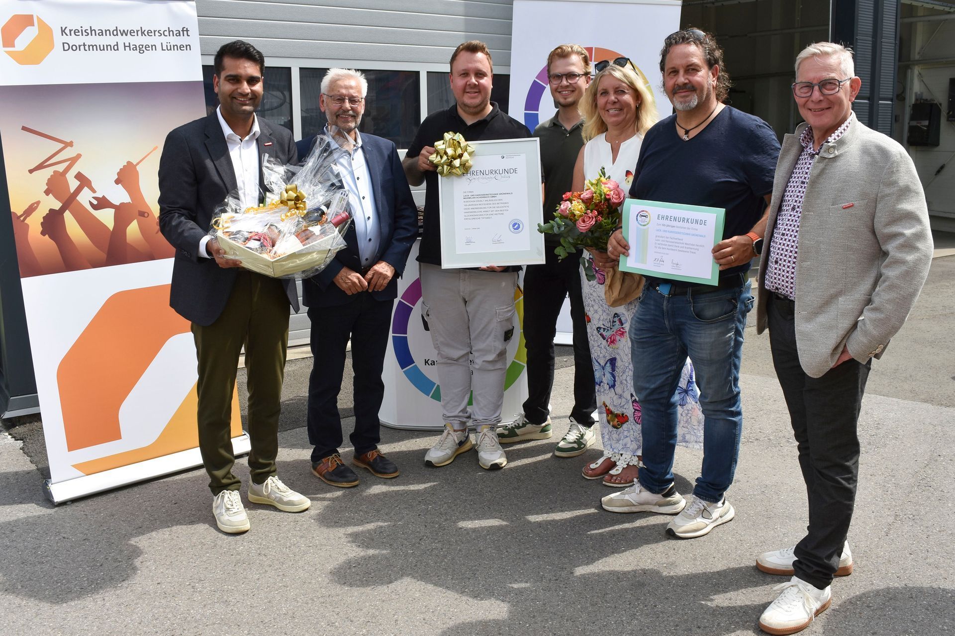 Eine Gruppe von Menschen posiert im Freien mit Blumensträußen und Urkunden bei einer Anerkennungszeremonie.
