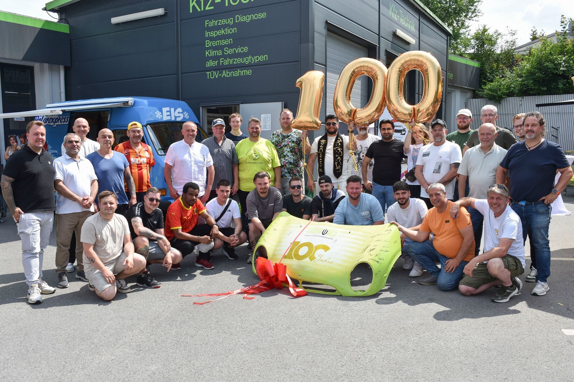 Eine Gruppe von Menschen posiert im Freien vor einem Gebäude und feiert mit einer großen goldenen „100“-Anzeige.