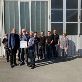 Eine Gruppe von Menschen steht vor einem Gebäude und hält im hellen Sonnenlicht eine Urkunde hoch.