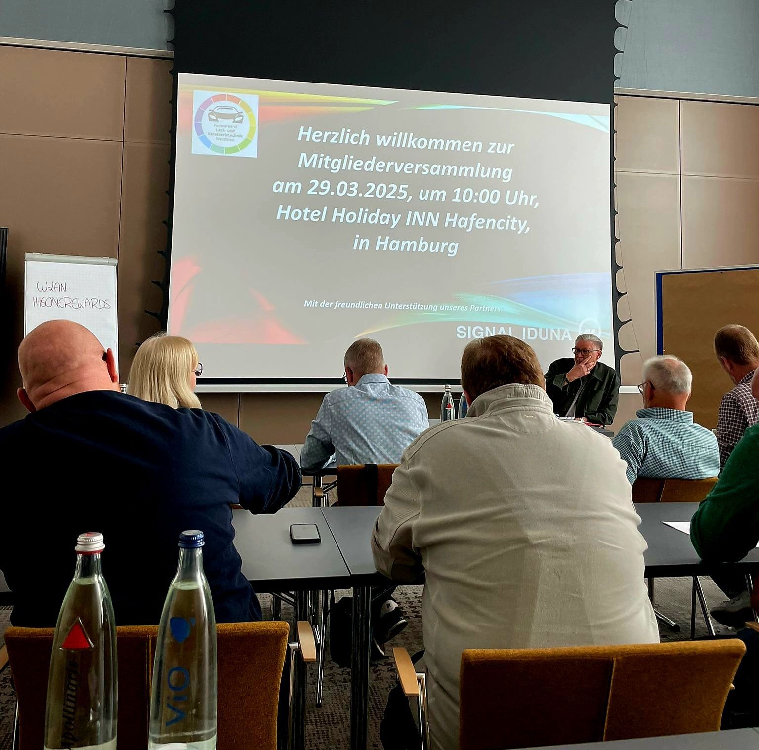 Publikum sitzt in einem Hotelkonferenzraum vor einer Projektionsfläche.