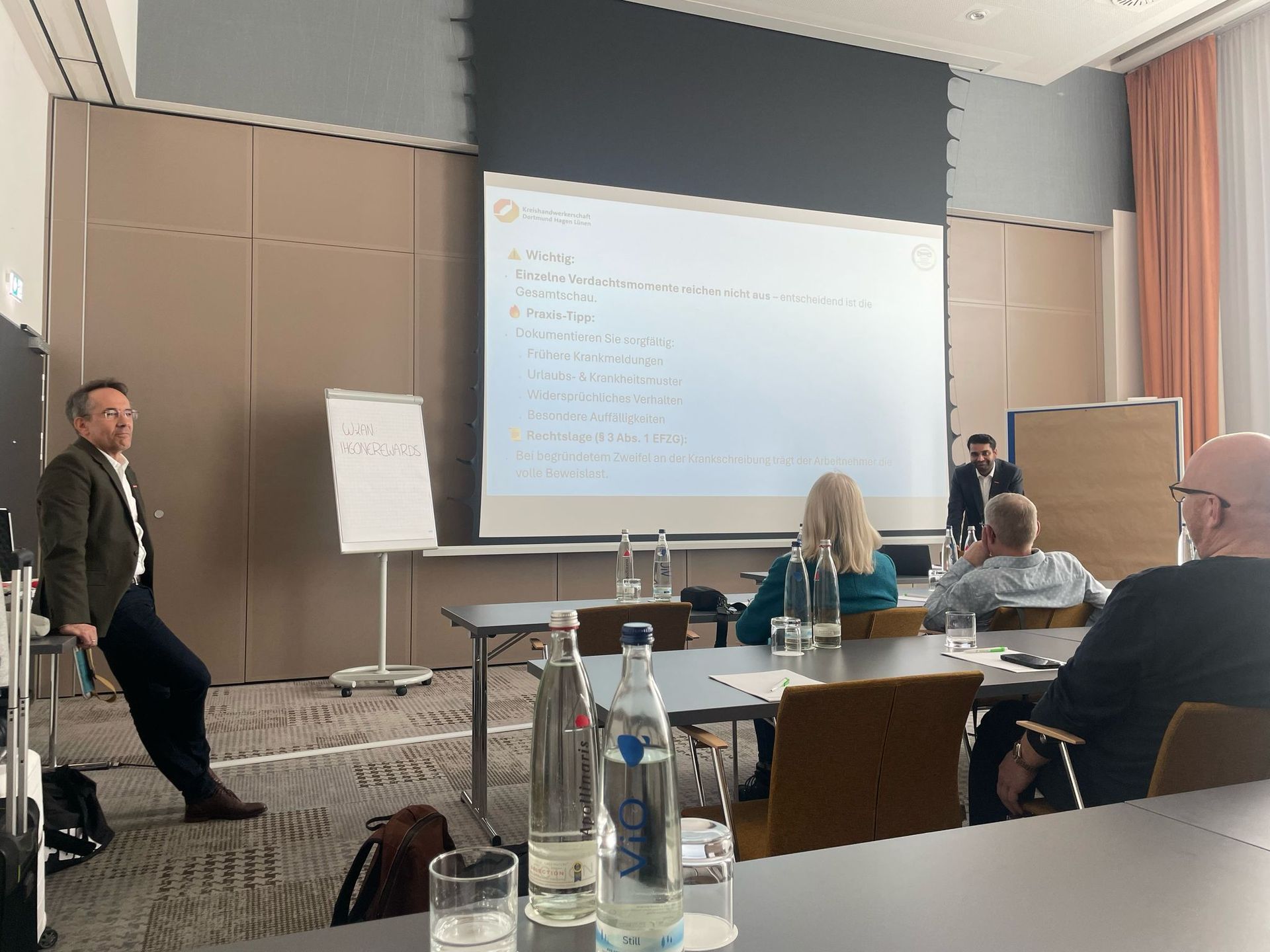 Präsentation im Konferenzraum mit projizierten Folien, sitzenden Teilnehmern und einem Präsentator neben einem Flipchart.