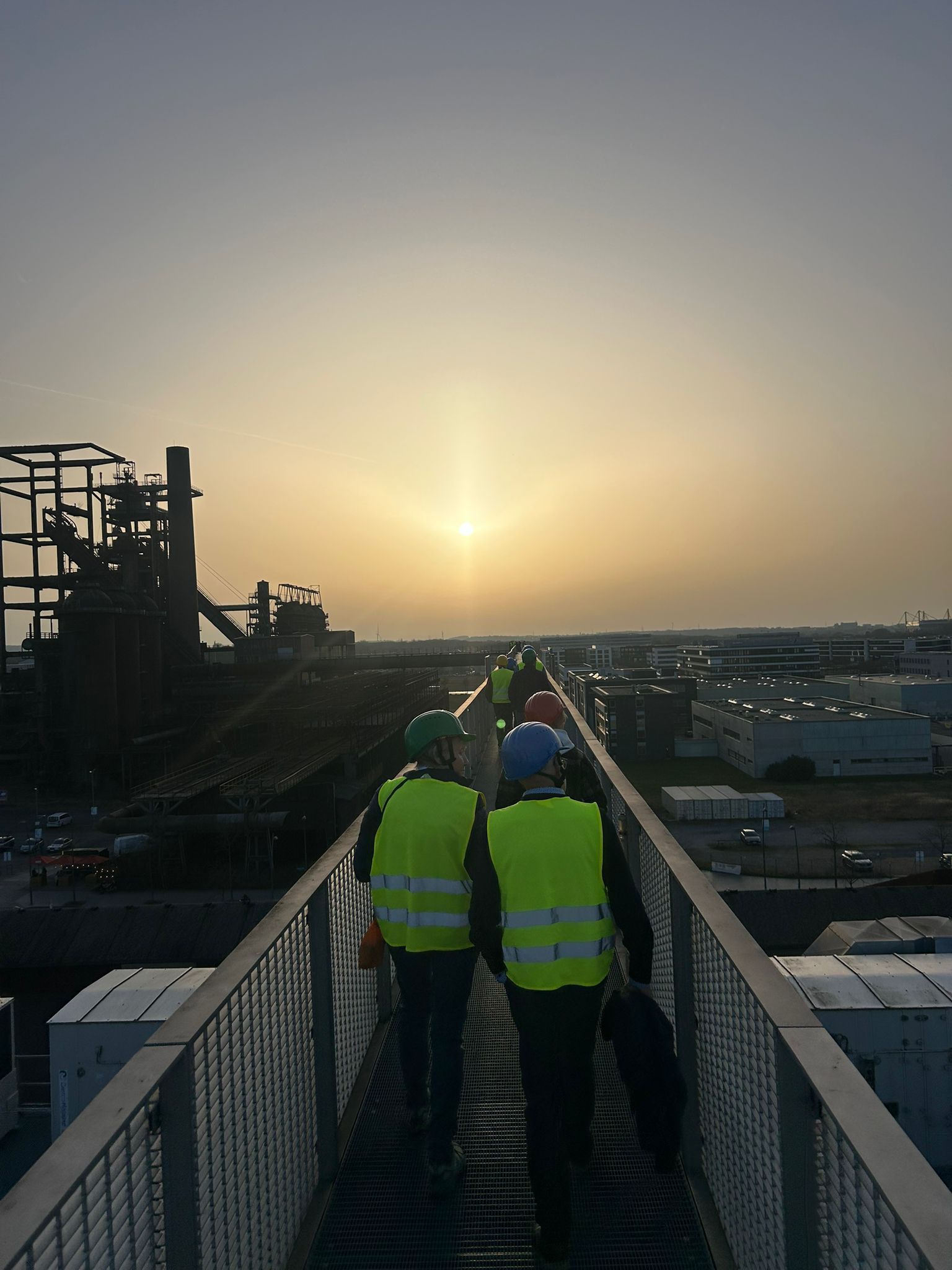 Arbeiter in neonfarbenen Sicherheitswesten gehen bei Sonnenuntergang auf einem erhöhten Industriesteg.