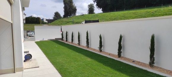 Lange, weisse Mauer mit mehreren schlanken Bäumen in einer Reihe. Grüner Rasen und ein Betonweg. Bewölkter Himmel.