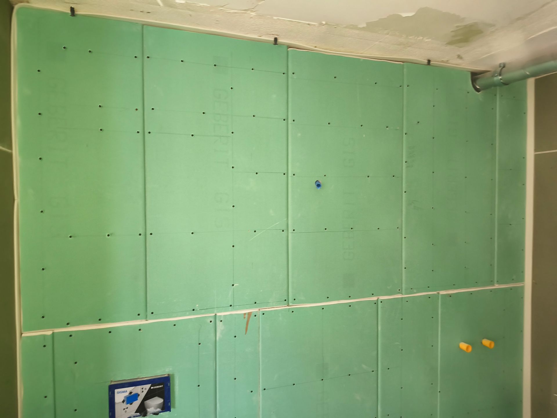 An einer Wand ist eine grüne Gipskartonplatte angebracht, die Schraubenköpfe sind sichtbar. Ein Rohr und elektrische Installationen sind ebenfalls vorhanden.