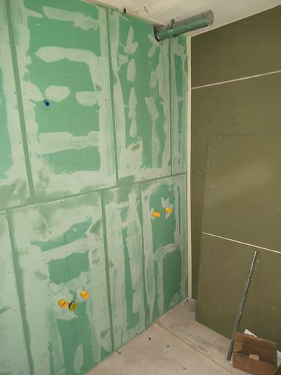 Innenwand mit grüner Gipskartonplatte, sichtbaren Sanitär- und Elektroverteilerkästen während der Bauphase.