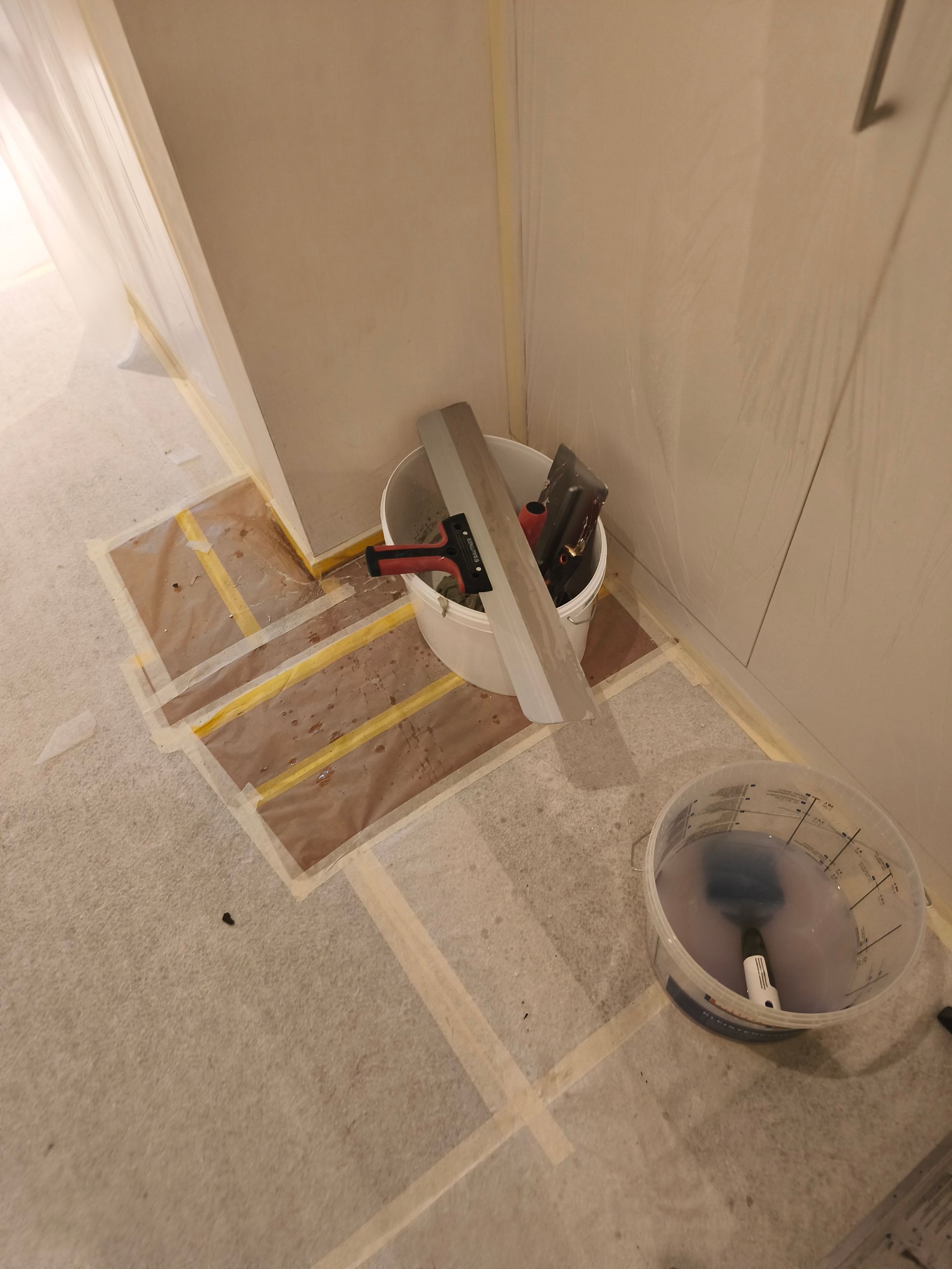 Bauwerkzeuge in Eimern neben einer Wand. Der Boden ist abgeklebt und mit Papier abgedeckt.