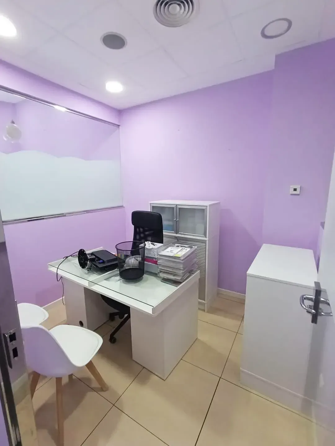 Oficina pequeña con paredes moradas, escritorio, sillas y armarios blancos. Se ven una pizarra blanca y material de oficina.