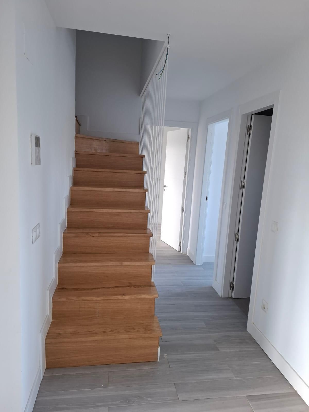 Escalera de madera que conduce al piso superior, pasillo con puertas abiertas, paredes y suelo de color claro.