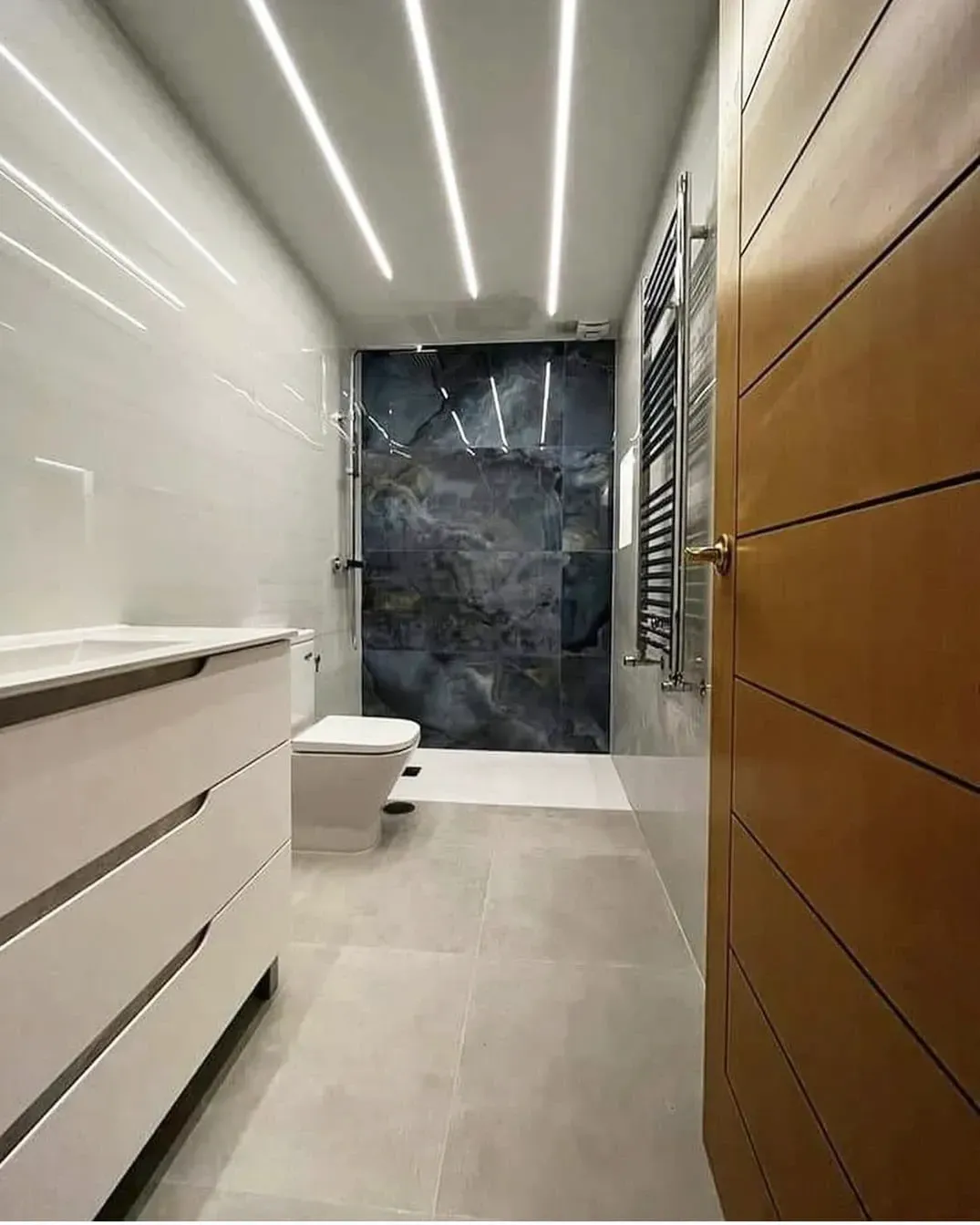 Baño moderno con azulejos blancos y grises, pared decorativa azul, tocador blanco y puerta de madera.