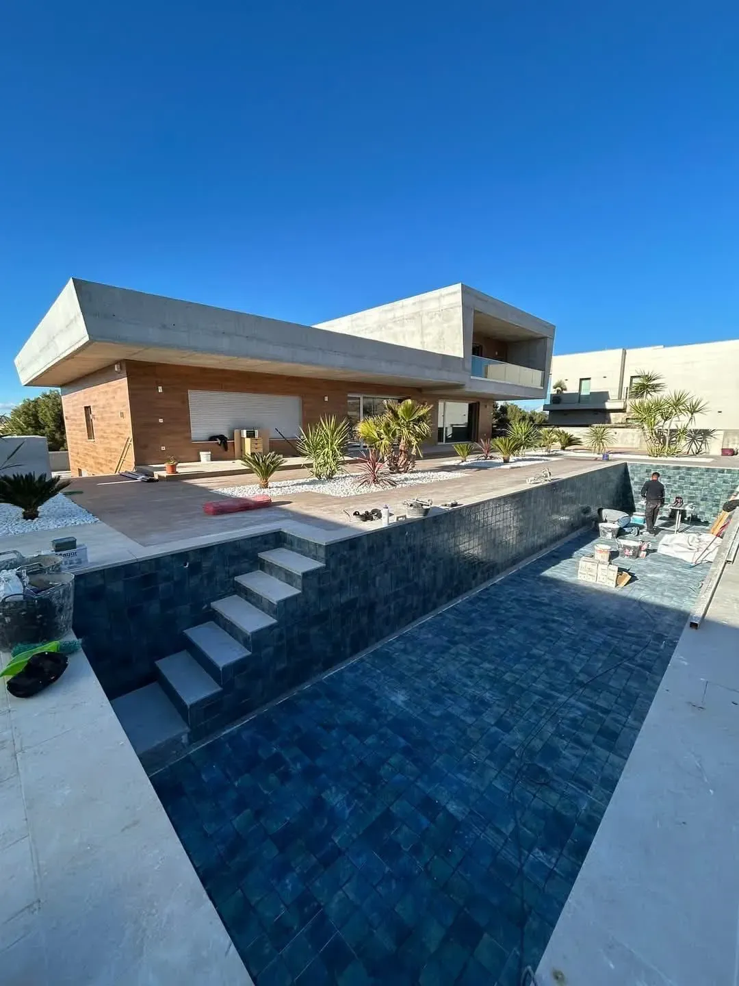 Casa moderna con piscina en construcción; azulejos azules en la piscina, escalones de hormigón y cielo azul.