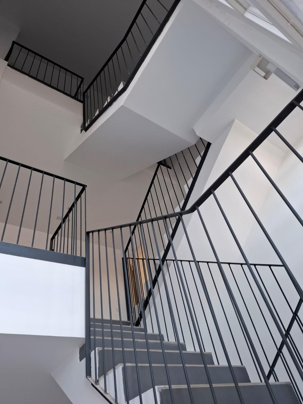 Vista interior de una escalera con barandillas de metal negro y peldaños grises. Las paredes son blancas.