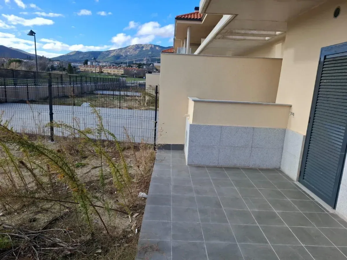 Patio con suelo de baldosas grises, paredes beige y vistas a una valla, vegetación y montañas bajo un cielo azul.