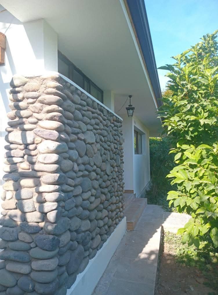 Muro revestido de piedra junto a un edificio blanco con una pasarela, vegetación verde a la derecha.