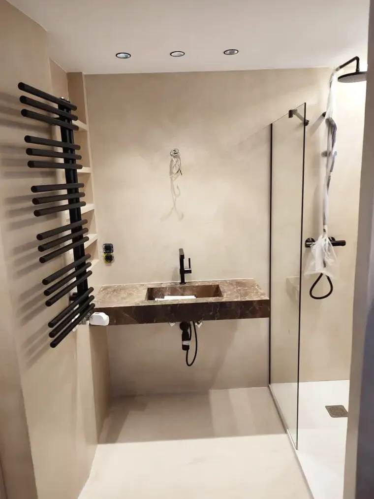 Baño moderno con lavabo de piedra marrón, ducha a ras de suelo y toallero negro. Paredes color beige.