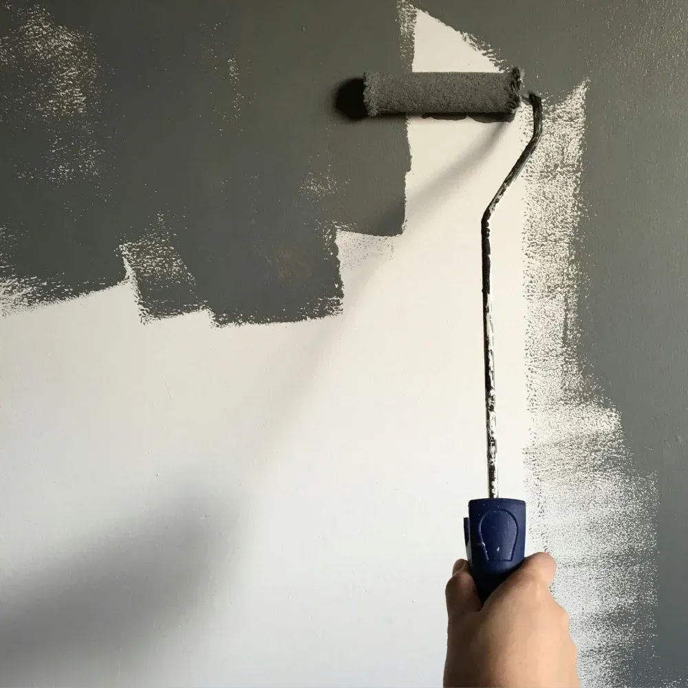 Una persona pinta una pared de gris con un rodillo; las secciones blancas y grises muestran el progreso.