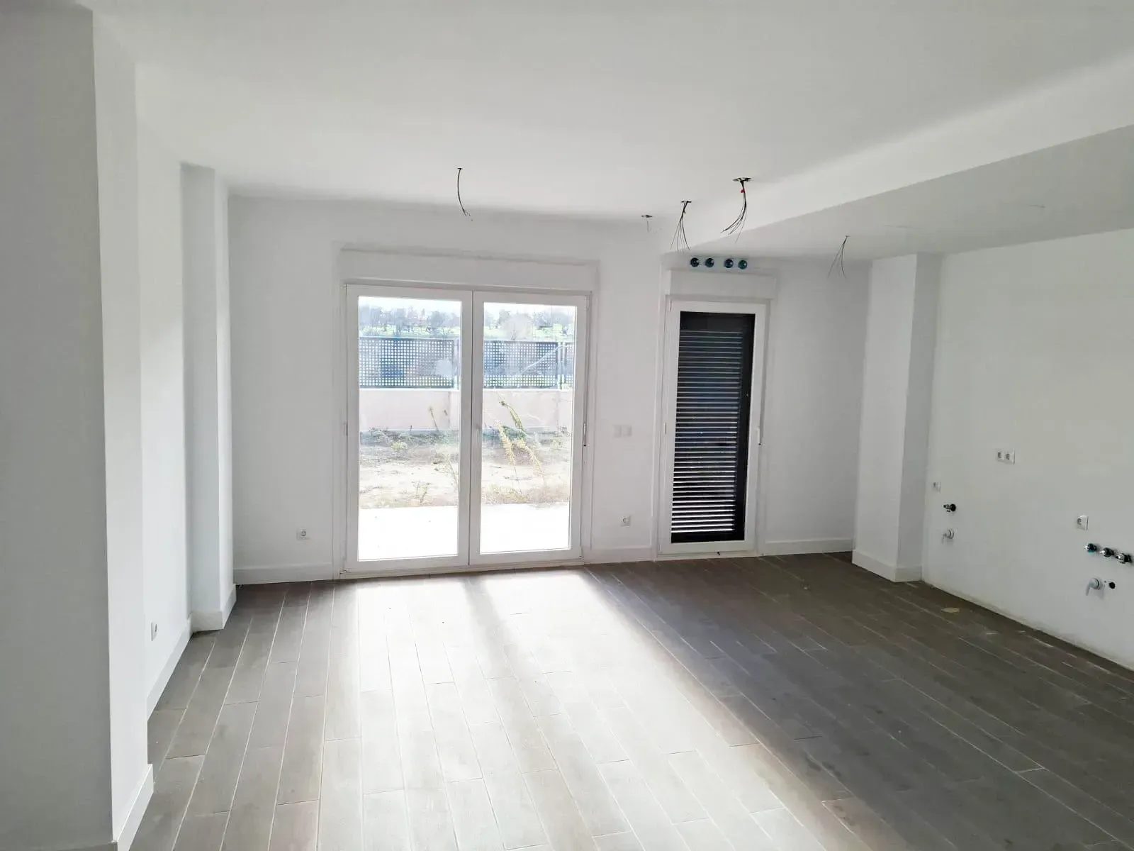 Habitación vacía con suelo de madera clara, paredes blancas y una puerta de cristal que da al exterior.