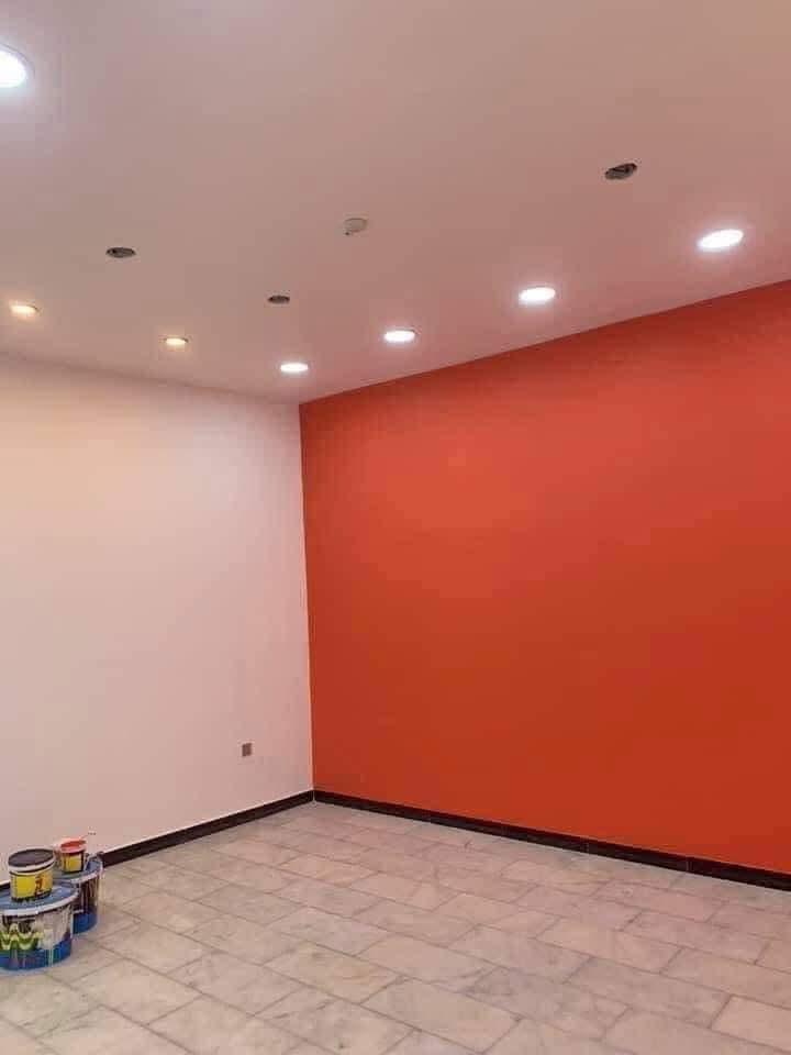 Habitación con paredes blancas y naranjas, iluminación empotrada y suelo de baldosas. Hay latas de pintura en el suelo.