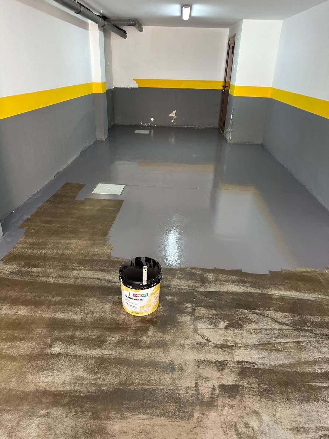 Garaje con suelo gris recién pintado, paredes amarillas y grises, cubo de pintura.