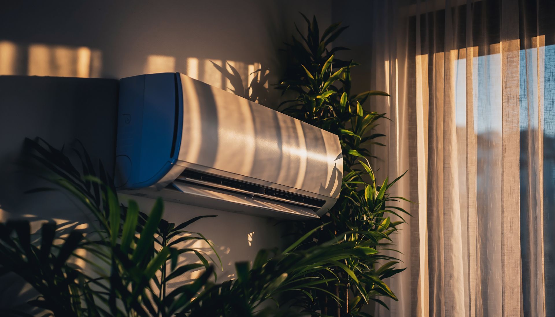 Aire acondicionado montado en una pared, rodeado de plantas, luz solar a través de una cortina transparente.