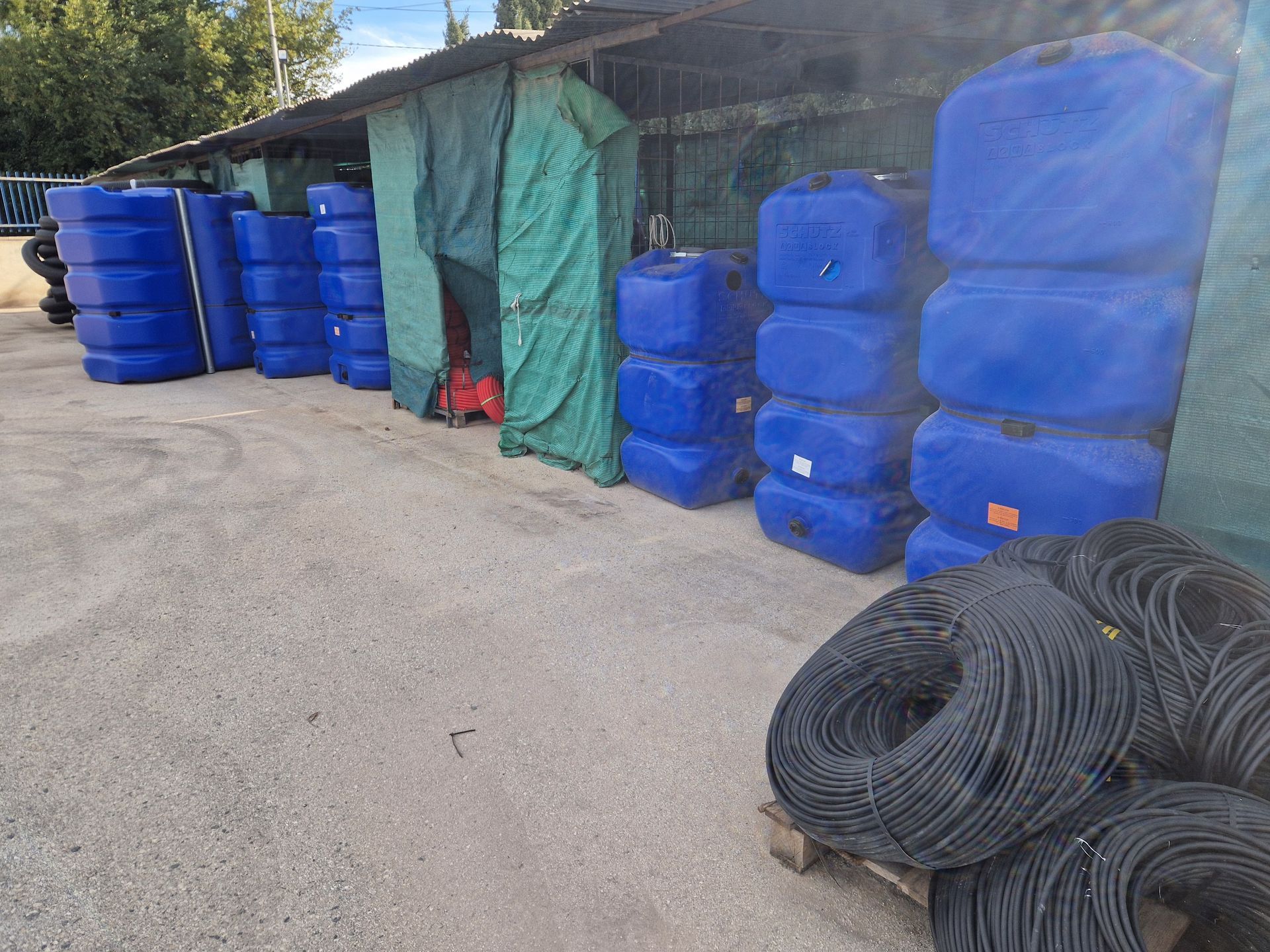 Tanques de plástico azul y carretes de alambre negro apilados afuera de un cobertizo.