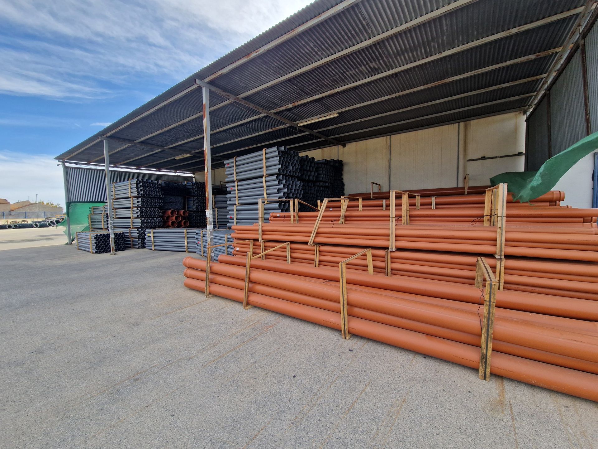 Filas de tubos naranjas y otros materiales almacenados bajo un techo de chapa ondulada. Cielo nublado al fondo.