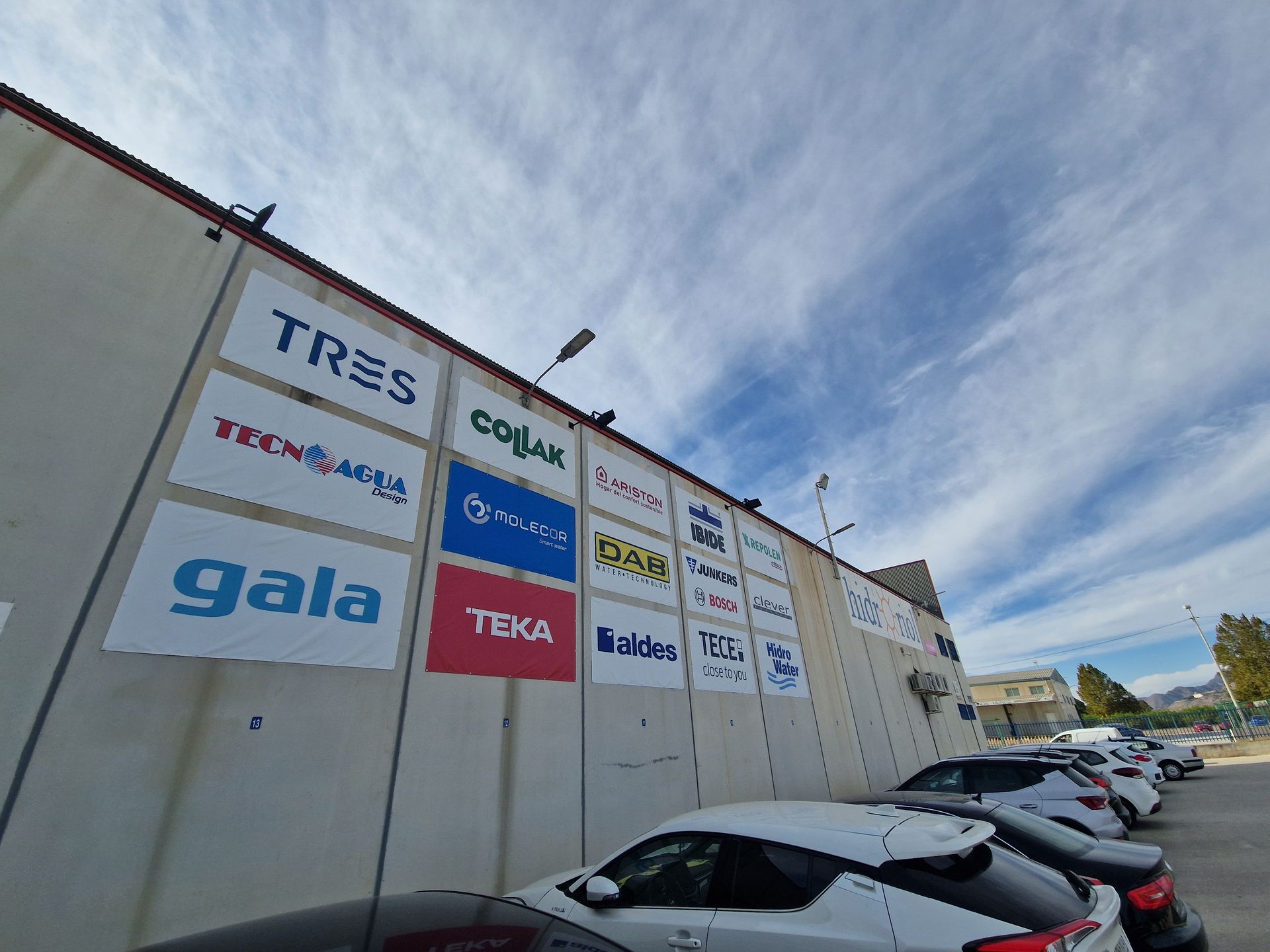 Muro con logotipos de empresas; coches aparcados debajo. Cielo azul con nubes.