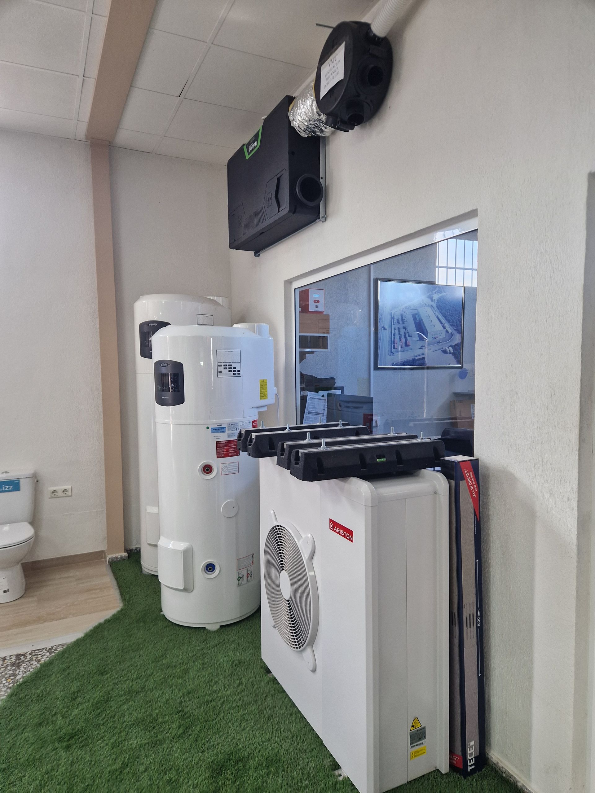 Unidades de calefacción y ventilación de color blanco expuestas en una sala de exposición, contra paredes blancas y césped artificial.