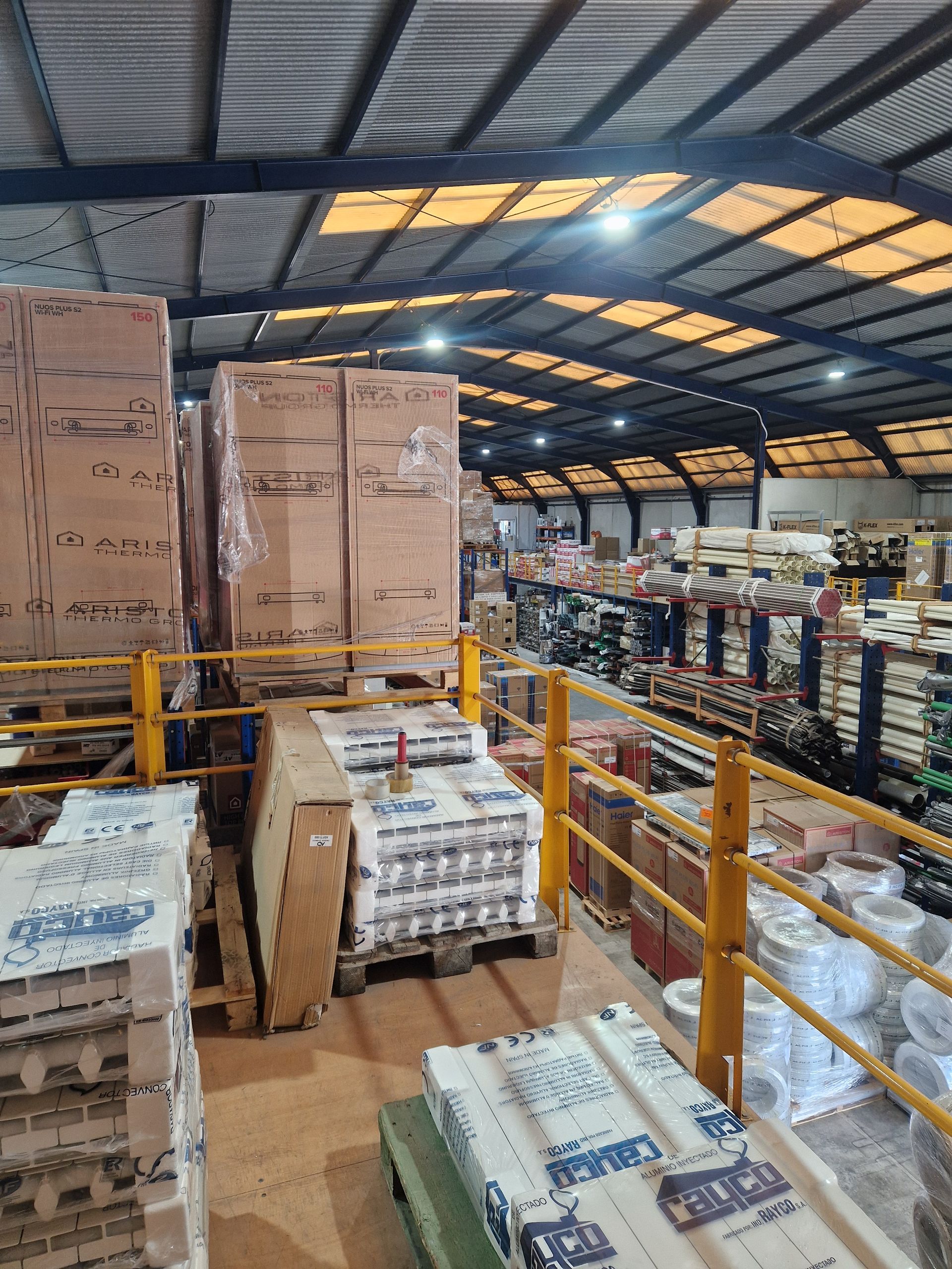 Interior de almacén con cajas y palés apilados. Barandilla de seguridad amarilla y mercancías en estanterías al fondo.