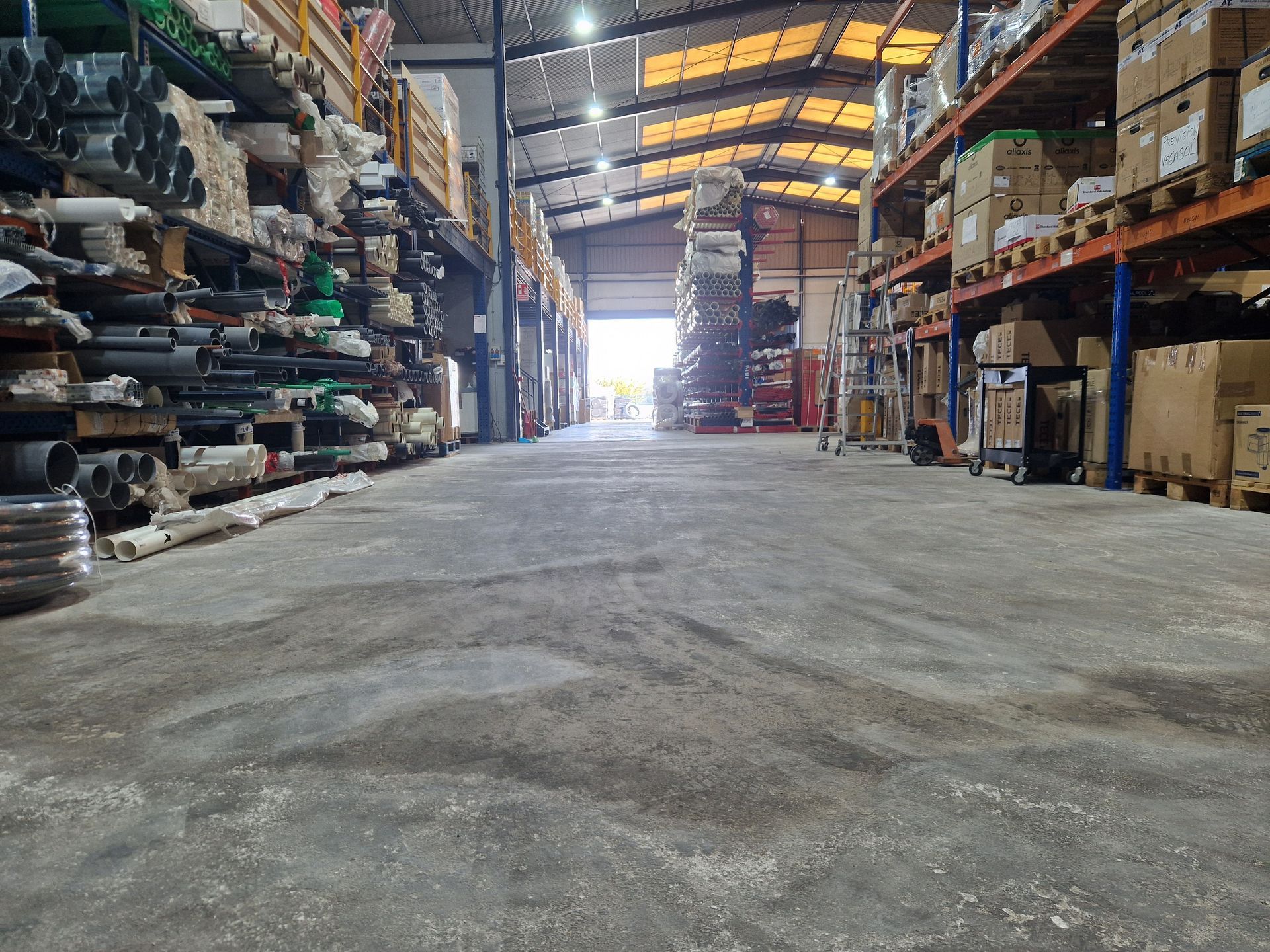 Interior de almacén, estantes llenos de materiales, luz natural brillante al final de un largo pasillo.