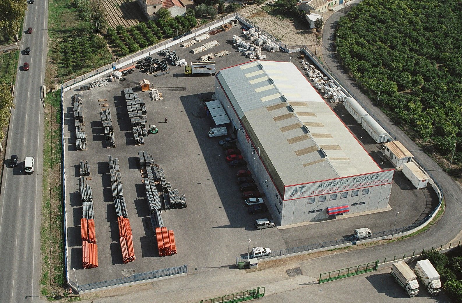 Vista aérea de un almacén y un patio. Diversos materiales industriales se almacenan en el exterior, rodeados de carreteras y zonas agrícolas.