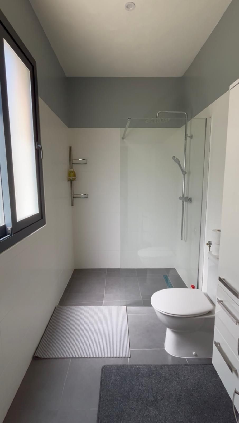 Salle de bains moderne avec douche à l'italienne, toilettes blanches, sol gris et lumière naturelle.