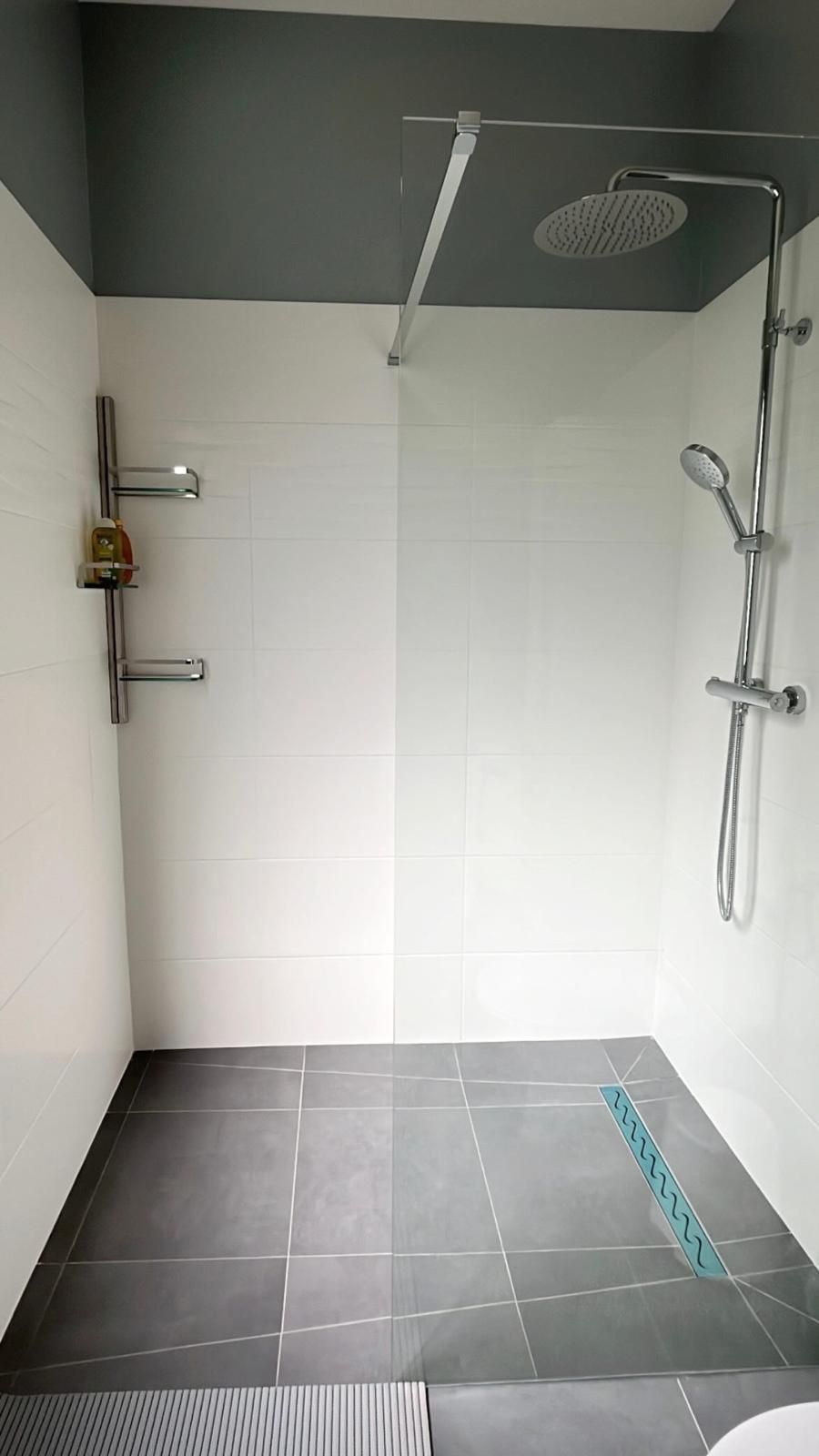 Douche accessible avec murs carrelés blancs, sol foncé, barres d'appui, douchette et pommeau de douche de plafond.