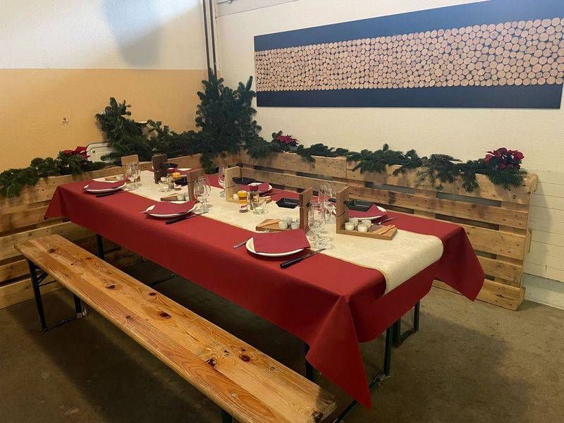 Festlich gedeckter Tisch mit roter Tischdecke, beigefarbenem Tischläufer und Holzbänken, geschmückt mit Grünpflanzen.