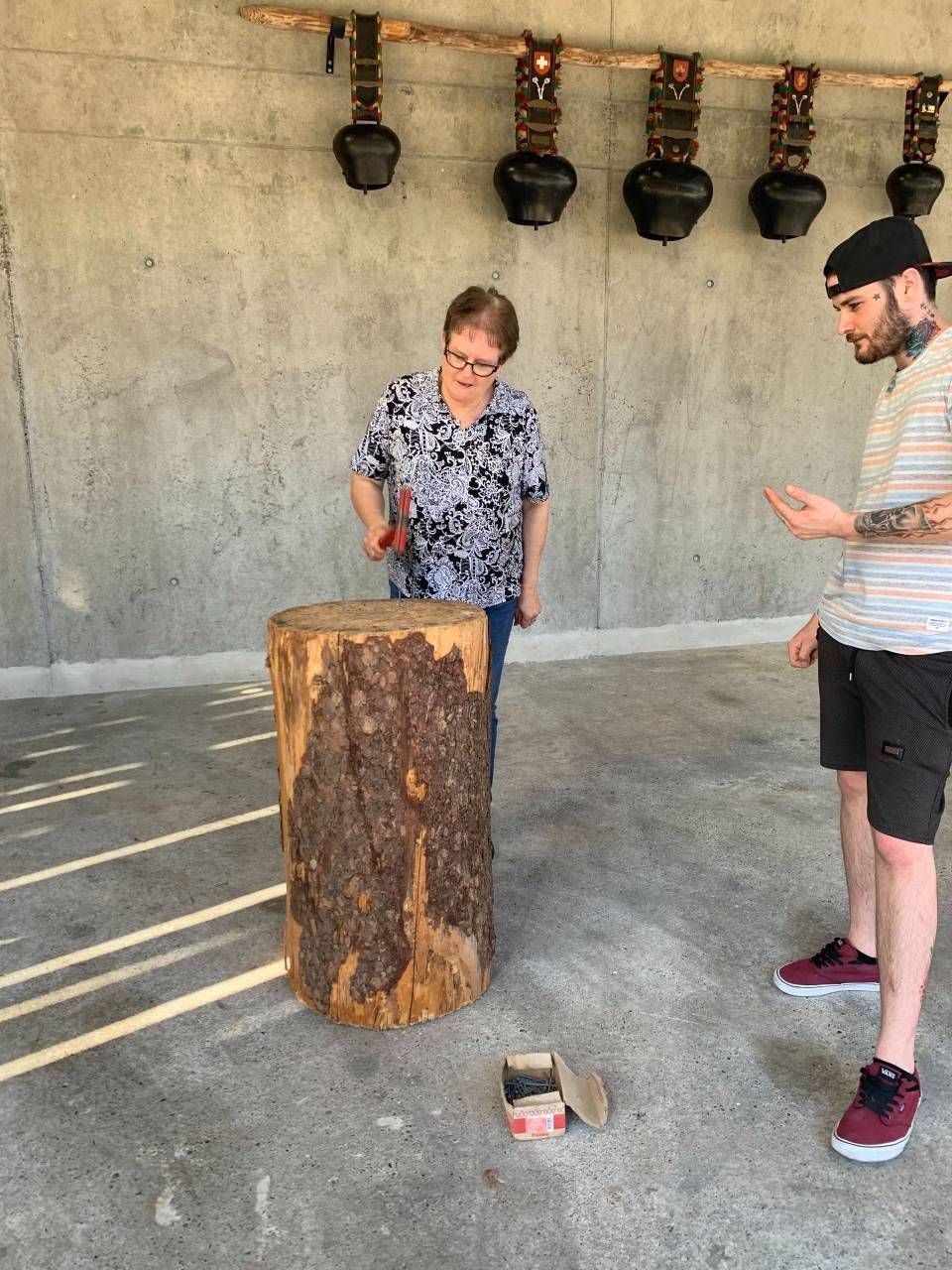 Eine Frau hämmert auf einen Holzstumpf, ein Mann schaut zu. Darüber hängen Glocken. Betonboden, sonnige Aussenkulisse.