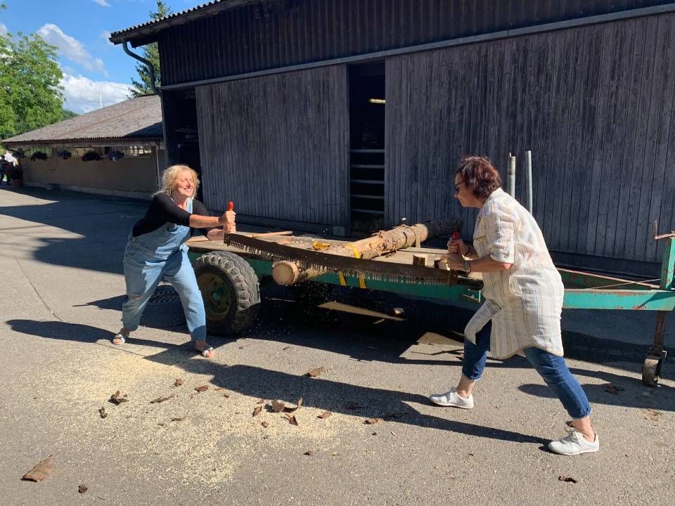 Zwei Personen sägen Holz auf einem Anhänger vor einem Holzgebäude. Sägemehl ist verstreut.